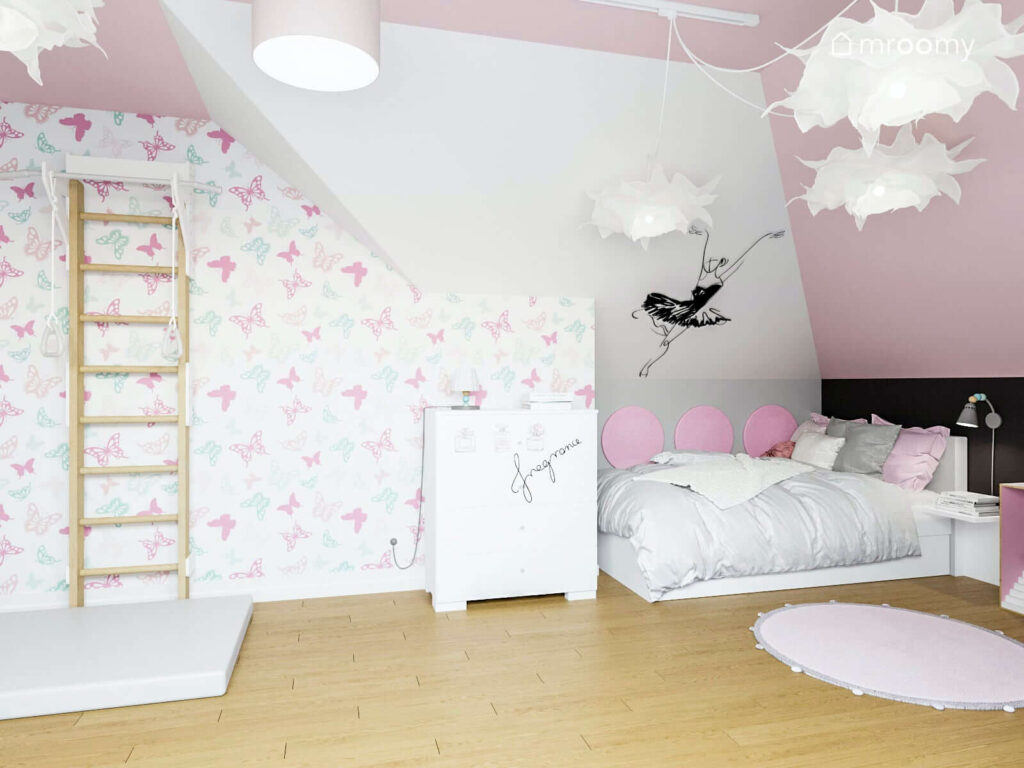 Drabinka gimnastyczna materac łóżko i komoda w biało różowym pokoju dziewczynki ozdobionym tapetą w motyle naklejką z baletnicą oraz miękkimi panelami ściennymi