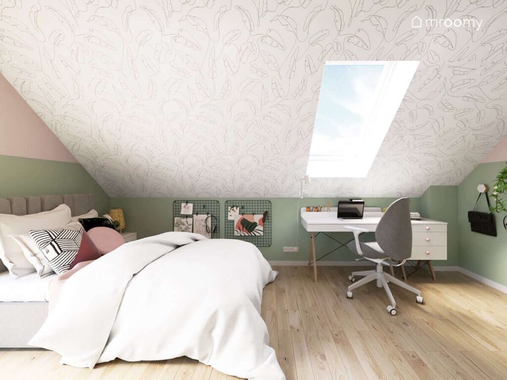 Skośna ściana z tapetą w liście oraz tapicerowane szare łóżko białe biurko i kontener a na ścianie organizery w poddaszowym pokoju dla dziewczynki