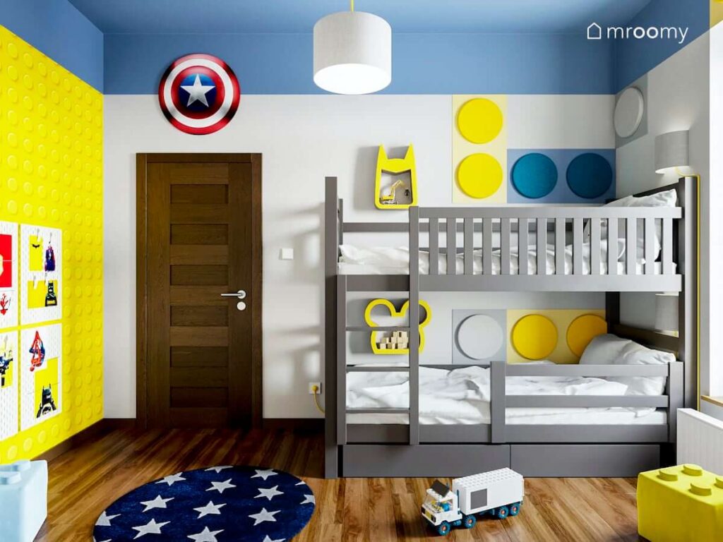 Piętrowe łóżko ozdobione kolorowymi miękkimi panelami ściennymi oraz półkami w żółto biało niebieskim pokoju dla rodzeństwa