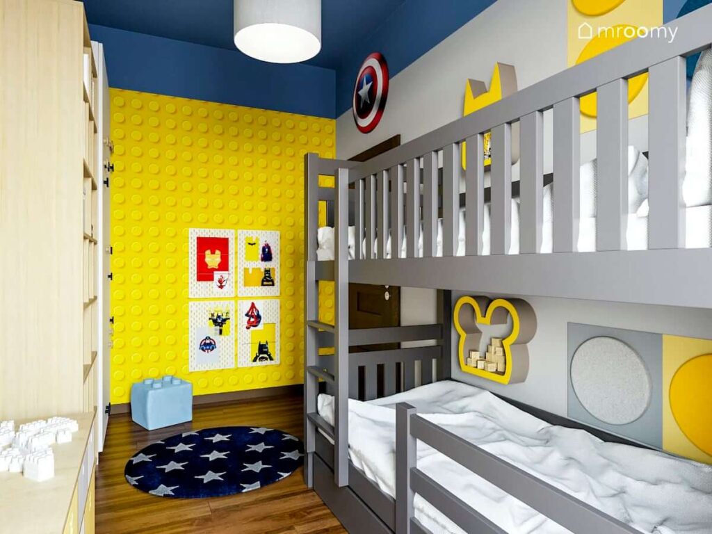 Żółto niebieski pokój z tapetą Lego dla rodzeństwa a w nim piętrowe łóżko oraz elementy nawiązujące do klocków Lego oraz superbohaterów