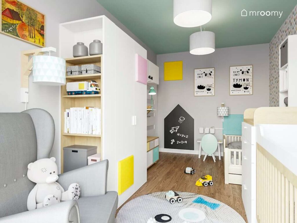 Pokoik rodzeństwa utrzymany w delikatnych pastelowych kolorach z dużą białą szafą drewnianymi łóżeczkami szarym fotelem i tablicą magnetyczną na ścianie