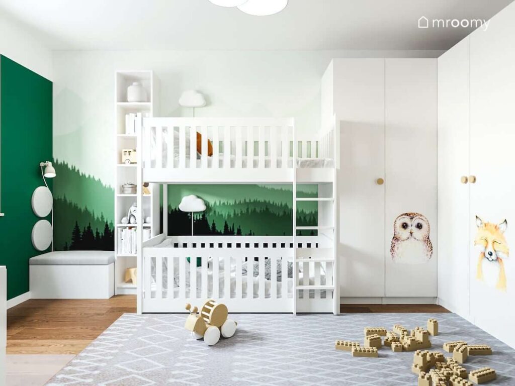 Tapeta przedstawiająca las oraz naklejki zwierzęta na dużej szafie w umeblowanym na biało pokoju dla rodzeństwa