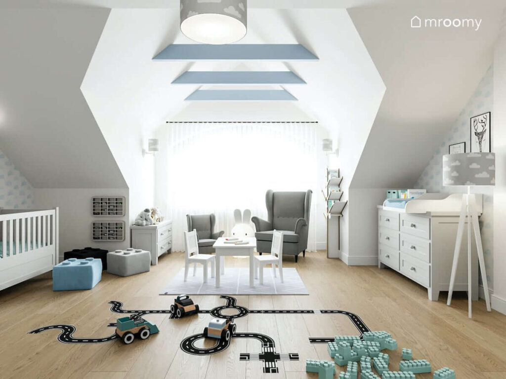 Duży jasny pokój na poddaszu dla rocznego chłopca z widocznymi drewnianymi belkami na suficie w kolorystyce szaro-błękitnej z półkami na resoraki i naklejkami podłogowymi w formie jezdni szarymi fotelami i dużym oknem