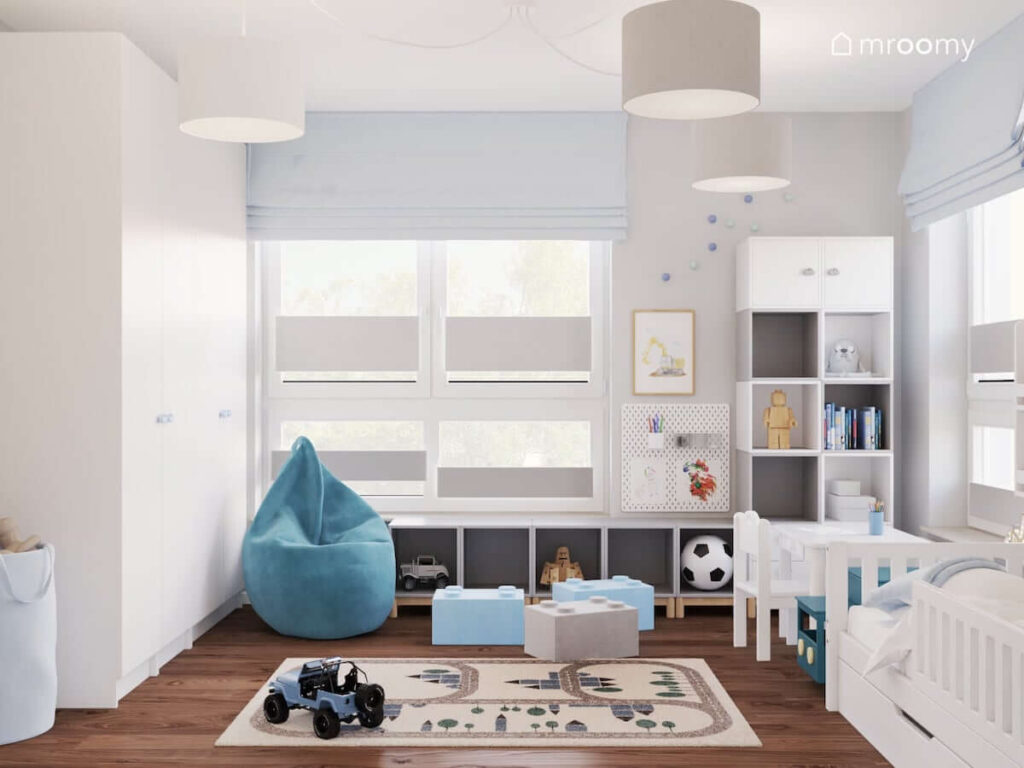 Jasny pokój dla chłopca z białymi meblami oraz niebieską pufą sako i małymi pufkami w kształcie klocków oraz dywanem z motywem miasteczka
