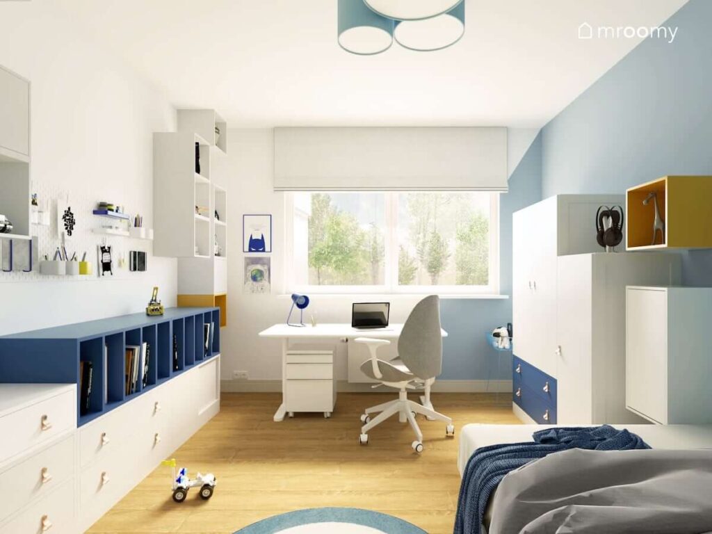 Białe szafki ścienne i organizery oraz biało niebieskie komody szafy i białe biurko pod oknem w pokoju dla chłopca