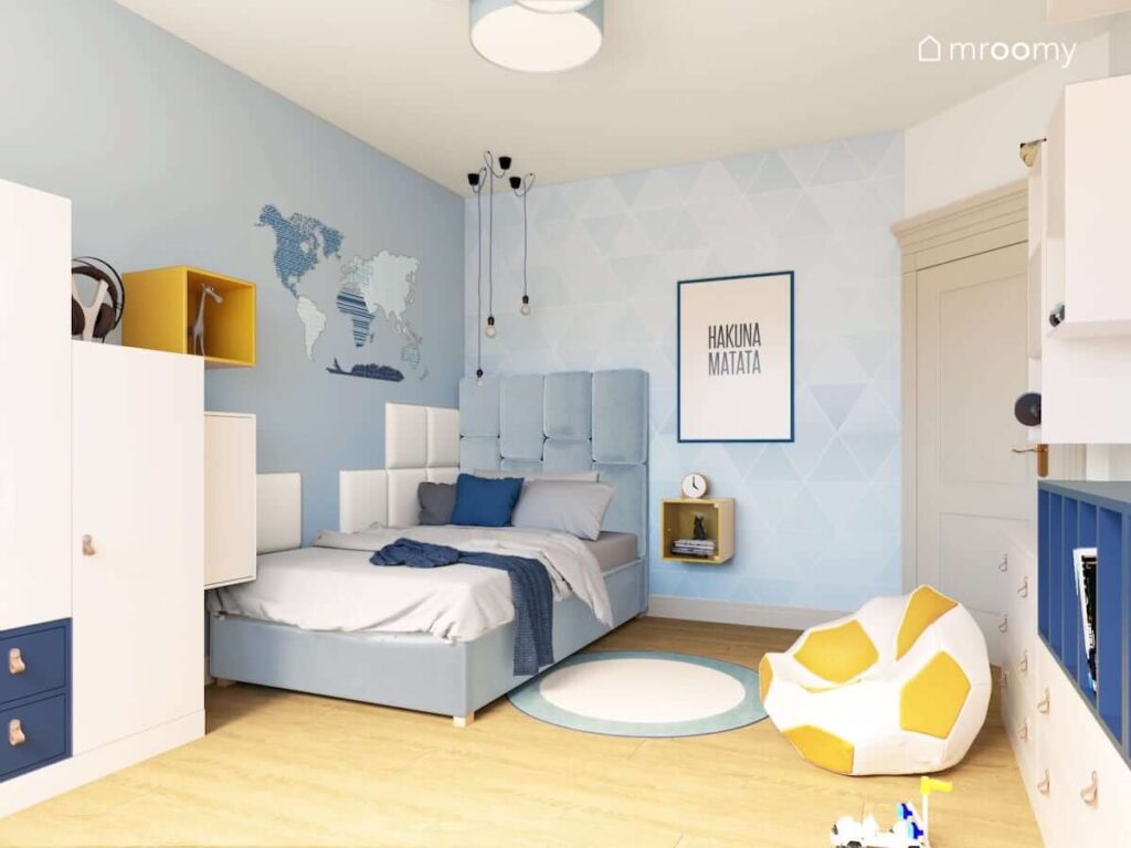 Pokój dla chłopca z niebieską tapetą w trójkąty oraz naklejką w kształcie mapy na ścianie oraz łóżkiem z wysokim niebieskim wezgłowiem i pufą w kształcie piłki