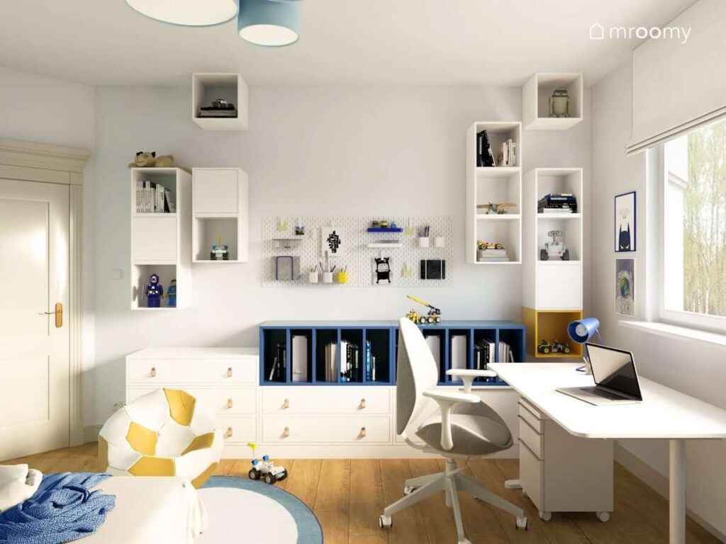 Liczne białe szafki ścienne biało niebieskie komody i organizery oraz białe biurko w pokoju dla chłopca