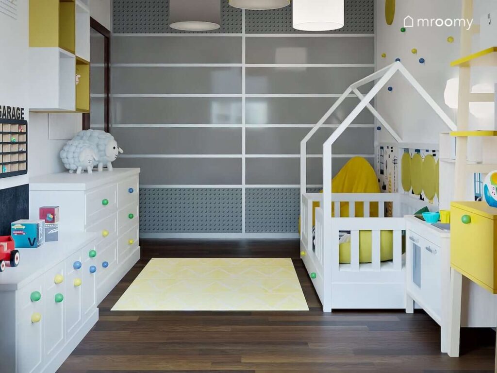 Pokój dla kilkuletniego chłopca z dużą szafą oklejoną szarymi naklejkami łóżkiem w kształcie domku i białymi komodami oraz półkami ściennymi w różnych kolorach