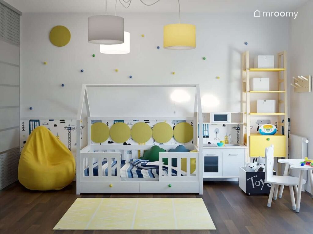 Jasny pokój dla chłopca z łóżkiem w kształcie domku żółtymi dodatkami oraz ścianą ozdobioną gałkami w różnych kolorach i tapetą z motywem budowy