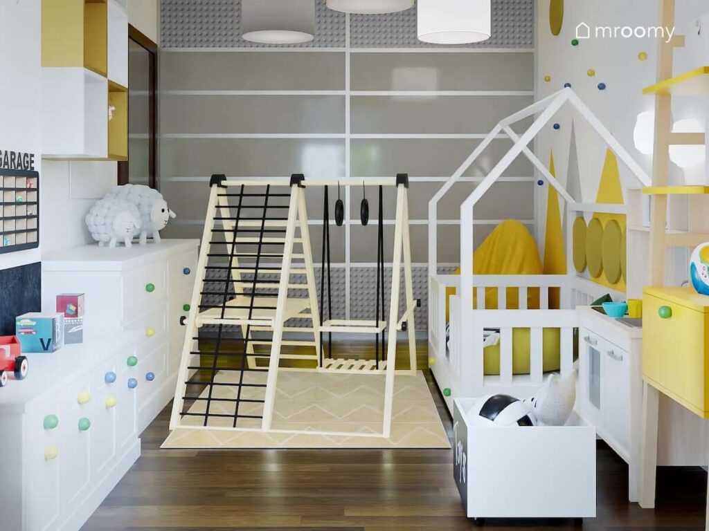 Drewniany plac zabaw oraz białe łóżko w kształcie domku w biało żółtym pokoju dla chłopca z białymi komodami