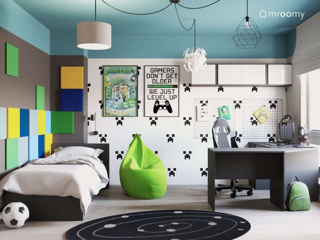 Pokój dla nastolatka fana gier komputerowych z wygodnym narożnym biurkiem okrągłym czarnym dywanem czarno-białą tapetą plakatem Minecraft zielonym workiem sako kwadratowymi piankowymi panelami na ścianie w kolorach żółtym błękitnym granatowym i zielonym lampą typu pajączek z różnymi abażurami