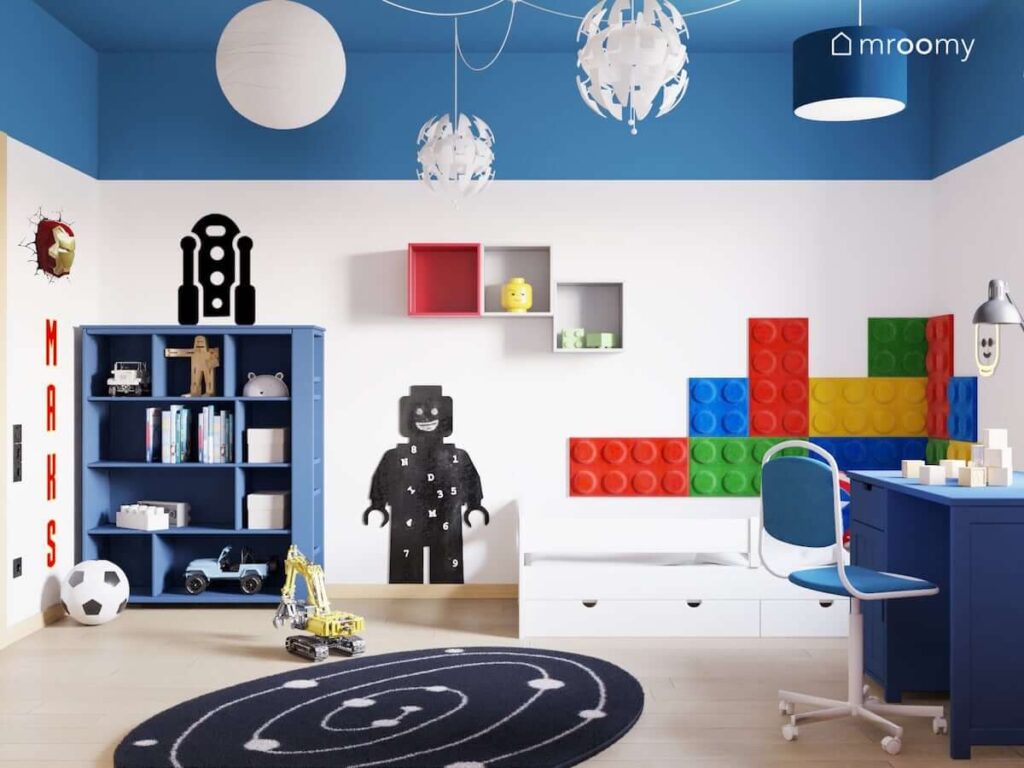 Pokój dla chłopca z kolorowymi panelami ściennymi w kształcie klocków zdobiącymi ściany przy łóżku a także z tablicą kredową w kształcie ludzika Lego i naklejką z R2D2 na ścianie i fantazyjnymi lampami sufitowymi na niebieskim suficie