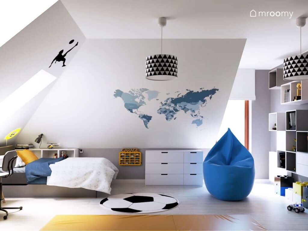 Poddaszowy pokój dla chłopca z łóżkiem półką na samochody niebieską pufą oraz skosami ozdobionymi naklejkami w kształcie piłkarzy i mapą świata