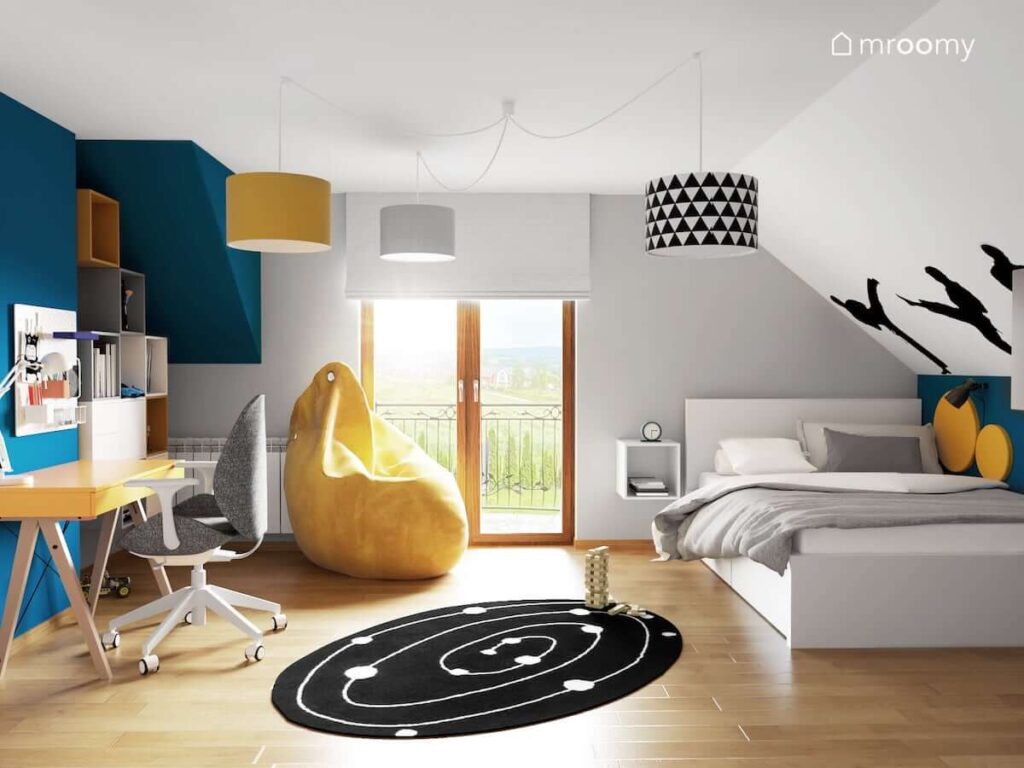Biało niebiesko żółty pokój na poddaszu dla chłopca z biurkiem pufą sako oraz dużym łóżkiem a także lampą sufitową z różnokolorowymi abażurami