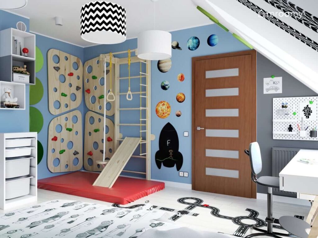 Ścianka wspinaczkowa oraz drabinka gimnastyczna ze zjeżdżalnią oraz tablica kredowa w kształcie rakiety i naklejki z planetami oraz biały organizer na ścianie pomalowanej farbą magnetyczną w pokoju kilkuletniego chłopca