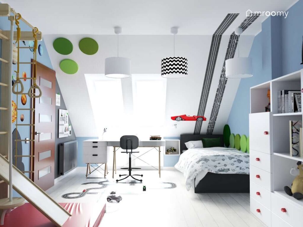 Poddaszowy pokój dla chłopca z białym biurkiem na drewnianych nogach pod oknem z łóżkiem oraz skosem ozdobionym zielonymi panelami ściennymi i naklejkami w kształcie śladów opon i wyścigówki