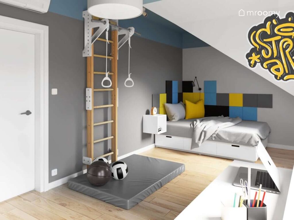Pokój dla chłopca z drabinką gimnastyczną szarym materacem gimnastycznym łóżkiem pod skosem kwadratowymi panelami ściennymi w kolorach żółtym czarnym i niebieskim z biurkiem skandynawskim pod oknem lampą z białym abażurem graffiti na ścianie