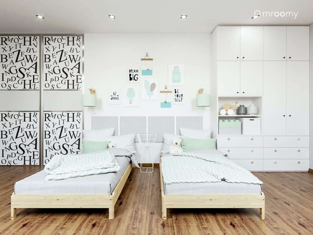 Dwa drewniane łóżka oraz duże szafy i ozdobne plakaty na ścianie obok białej szafy z kolorowymi uchwytami w pokoju dla dwóch dziewczynek