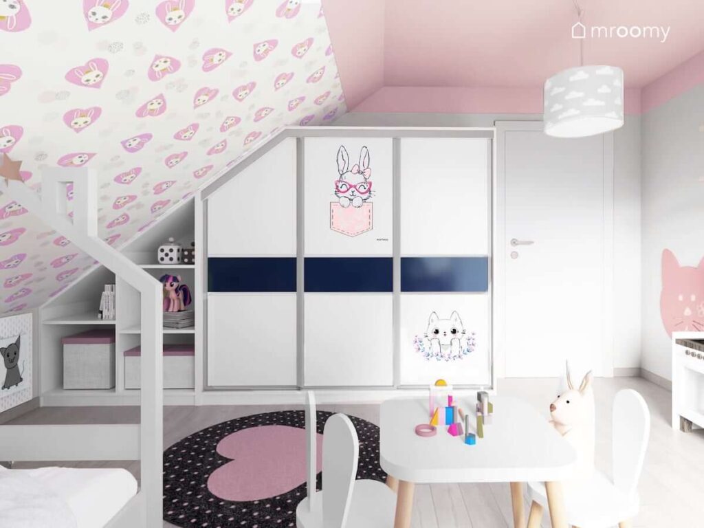 Dziewczęcy pokój na poddaszu z tapetą w serduszka i króliki różowym sufitem i duża szafą pod skosem obok łóżko domek