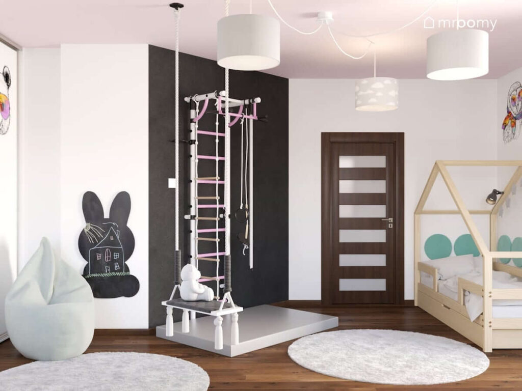 Jasny pokój dla dziewczynki z drabinką gimnastyczną umocowaną na czarnej ścianie oraz tablicą kredową w kształcie królika i drewnianym łóżkiem w kształcie daszku oraz huśtawką