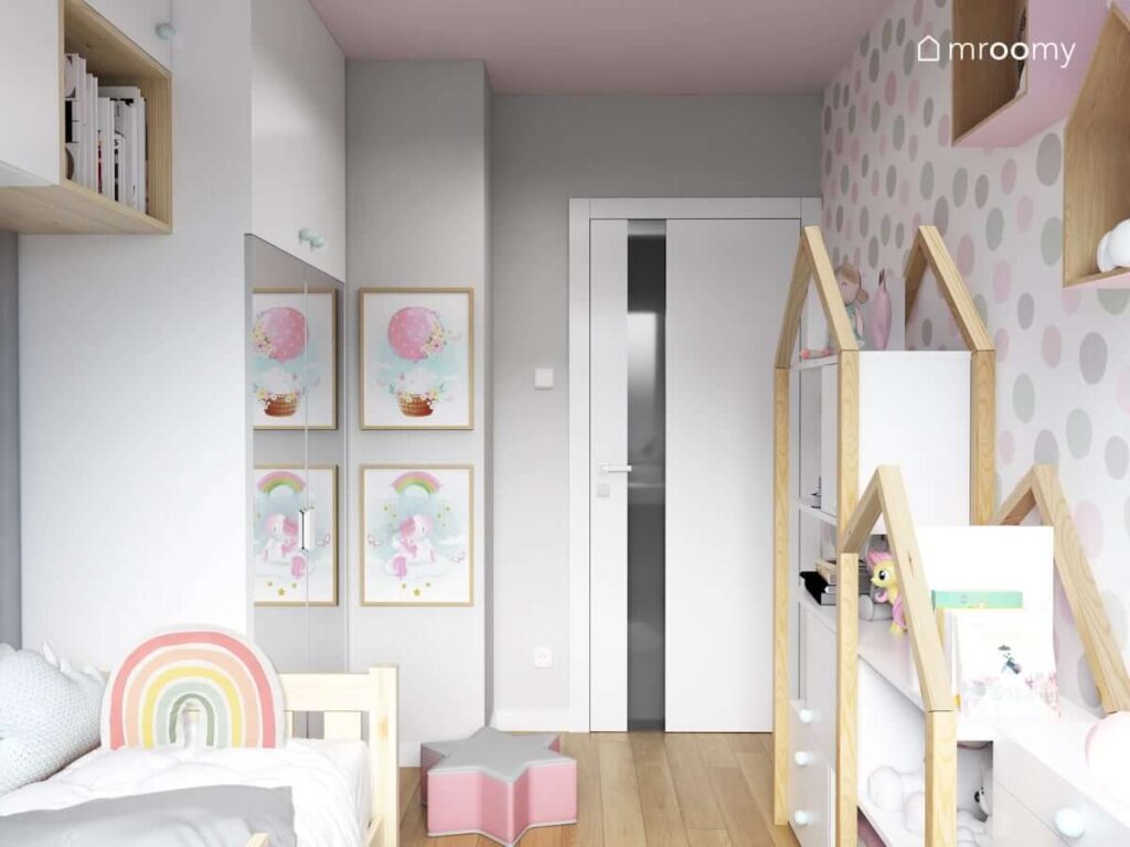 Wąski pokój dla dziewczynki w szaro-różowej kolorystyce z białymi meblami z elementami drewna naturalnego z poduszką w kształcie tęczy i pufą w kształcie gwiazdy z tapetą w szaro-różowe kropki