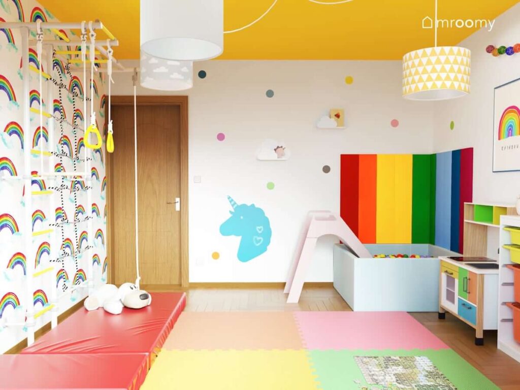 Kolorowy pokój dla dziewczynki z drabinką gimnastyczną basenem z kulkami i zjeżdżalnią z tablicą magnetyczną w kształcie jednorożca i tapetą z  motywem tęczy na ścianie kolorowymi panelami piankowymi na ścianie i kolorowymi puzzlami piankowymi na podłodze