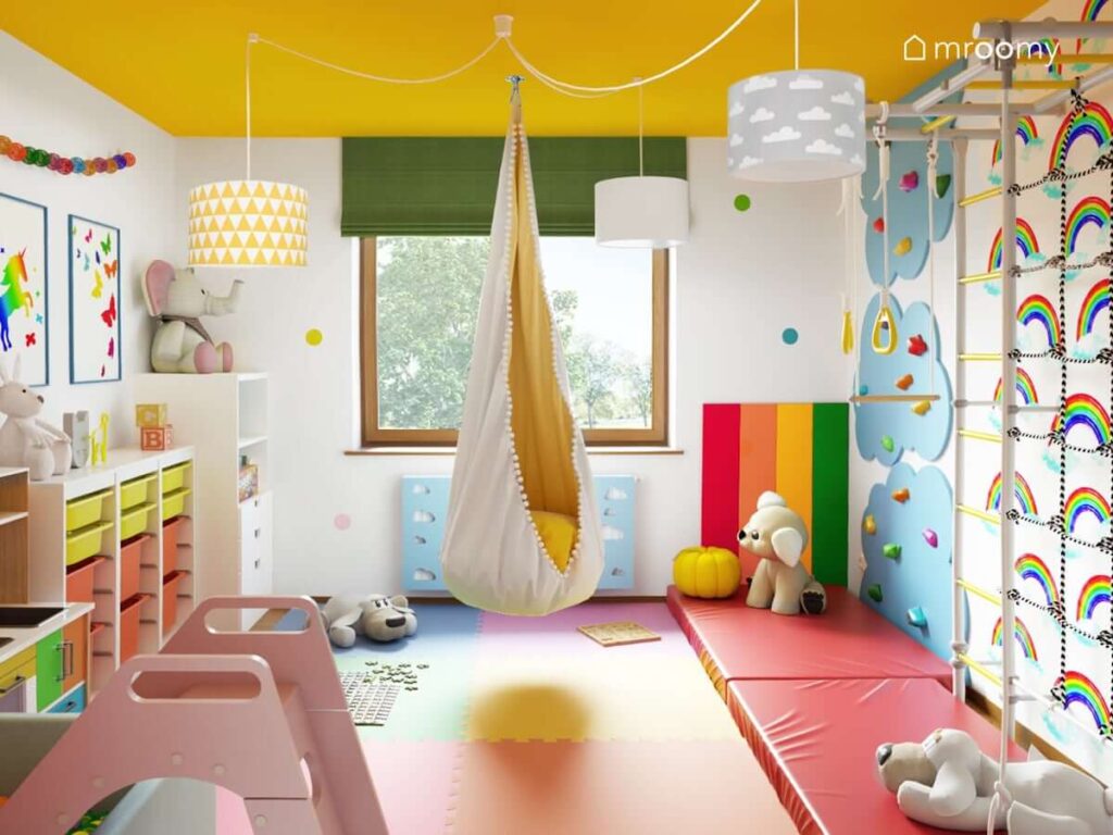 Kolorowy pokój zabaw ze ścianką do wspinaczki huśtawką basenem z kulkami i różową zjeżdżalnią piankowymi panelami ściennymi w kolorach tęczy żółtym sufitem i zieloną roletą rzymską