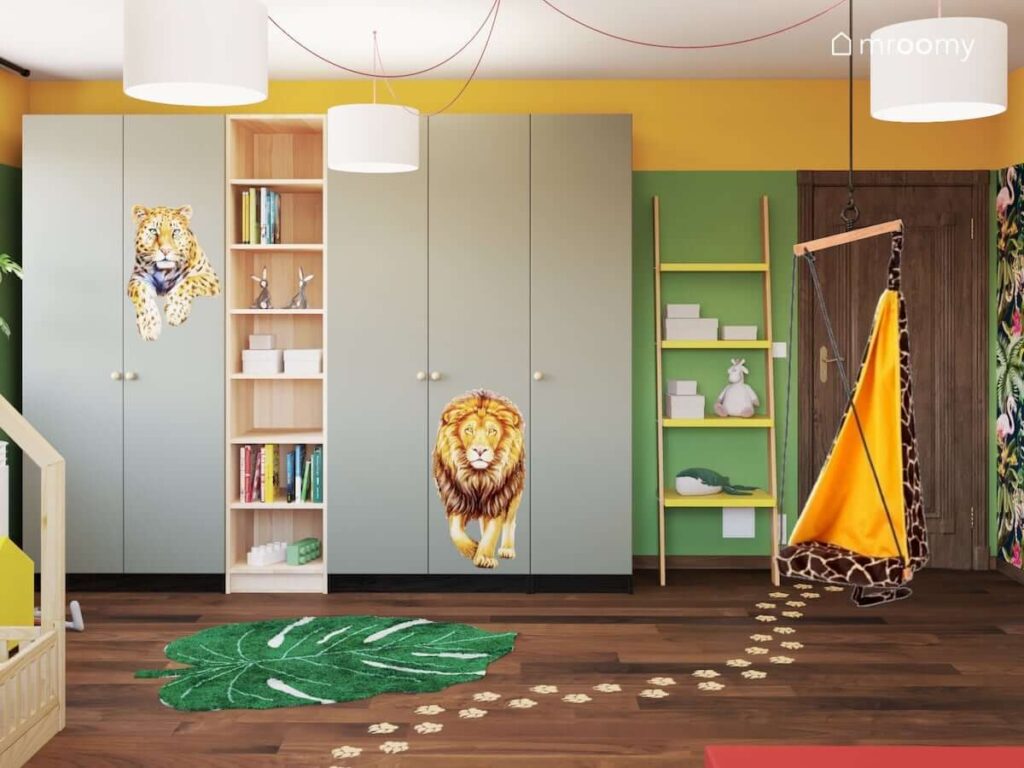 Szafy ozdobione naklejkami z dzikimi kotami oraz regał w kształcie drabinki a na podłodze dywan w kształcie liścia i naklejki połogowe w kształcie odcisków łap w zielono żółtym pokoju dla dziewczynki