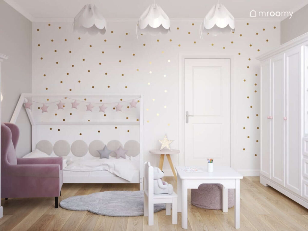 Biała ściana w złote groszki łóżko w kształcie domku udekorowane girlandą gwiezdną biały stolik z krzesełkiem oraz różowy fotel a na suficie białe lampy w kształcie bezy w jasnym pokoju dla małej dziewczynki