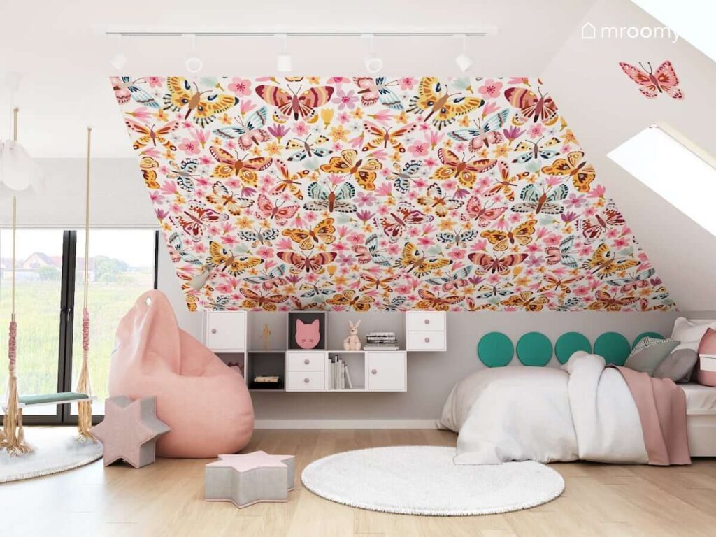 Pokój dla dziewczynki z dużą różową pufą sako białymi dywanami oraz kombinacją biało szarych szafek ściennych oraz ścianą pokrytą tapetą w motyle