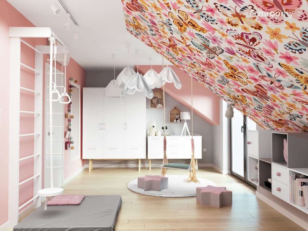 Biało-różowy pokój na poddaszu dla dziewczynki z białymi meblami wiszącą huśtawką małymi pufkami w kształcie gwiazdek oraz skosem oklejonym tapetą w motyle