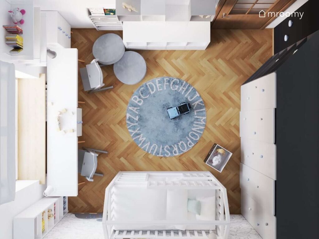 Kwadratowy pokój dziecka w kolorystyce biało-błękitnej z szarymi dodatkami z łóżkiem piętrowym i niebieskim okrągłym dywanem z alfabetem z dużym biurkiem pod oknem