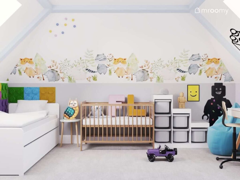 Białe łóżko i drewniane łóżeczko dziecięce oraz regał z pojemnikami na zabawki a także ściana ozdobiona naklejką z leśnymi zwierzątkami w pokoju dla dwóch sióstr