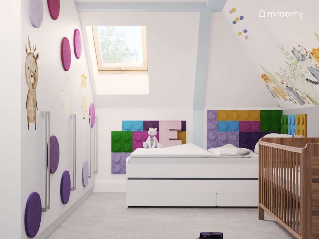 Jasny pokój dla rodzeństwa ze ścianami i frontami szaf ozdobionymi kolorowymi panelami ściennymi w różnych kształtach a także białym łóżkiem i drewnianym łóżeczkiem dziecięcym