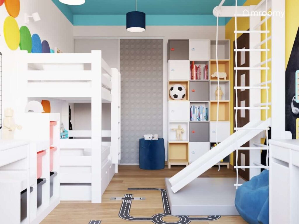 Biało niebiesko żółty pokój dla rodzeństwa z łóżkiem piętrowym licznymi szafkami ściennymi oraz szafą ozdobioną naklejką przypominającą wzór na klockach Lego