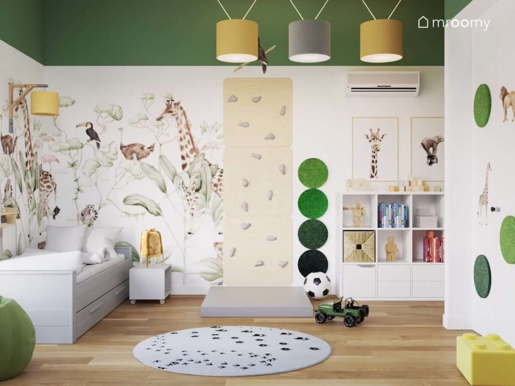 Ściana ozdobiona tapetą oraz plakatami z dzikimi zwierzętami oraz zielonymi panelami ściennymi a także łóżko z szufladą ścianka wspinaczkowa biały regał i dywan w łapki a na suficie żółte i szare lampy w pokoju dla przedszkolaka