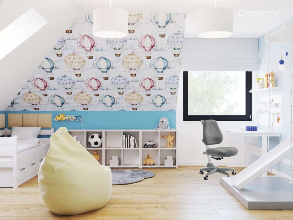 Ściana ozdobiona tapetą w balony oraz niskie bielone regały i łóżko a także biurko a na suficie białe lampy w pokoju dla chłopca