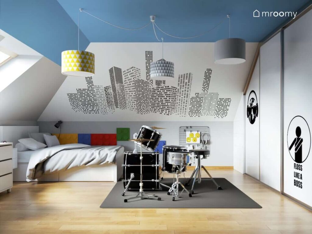 Biały pokój dla chłopca z niebieskim sufitem i perkusją na środku a na ścianie tapeta z graficznymi wieżowcami oraz panele piankowe w kształcie klocków