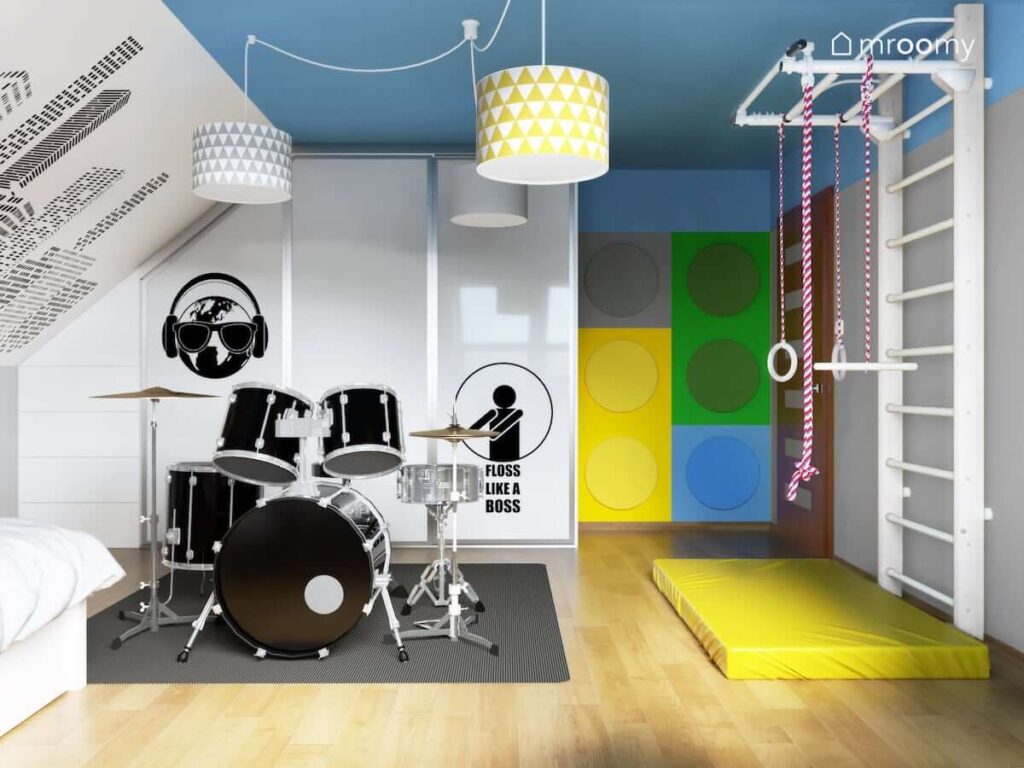 Pokój dla chłopca perkusisty z białą szafą ozdobioną naklejkami drabinką gimnastyczną z materacem oraz perkusją a na ścianie miękkie panele ścienne przypominające klocki Lego
