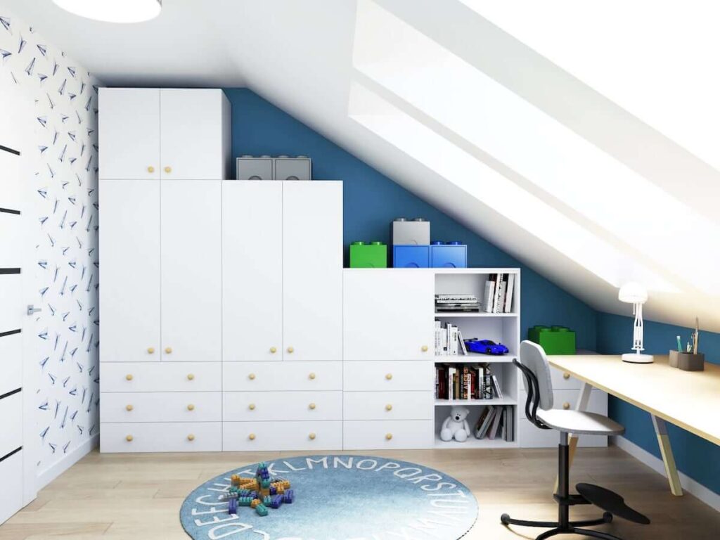 Biała szafa modułowa z drewnianymi gałkami oraz drewniane biurko pod skosem a na podłodze niebieski dywan z alfabetem w biało niebieskim poddaszowym pokoju dla chłopca
