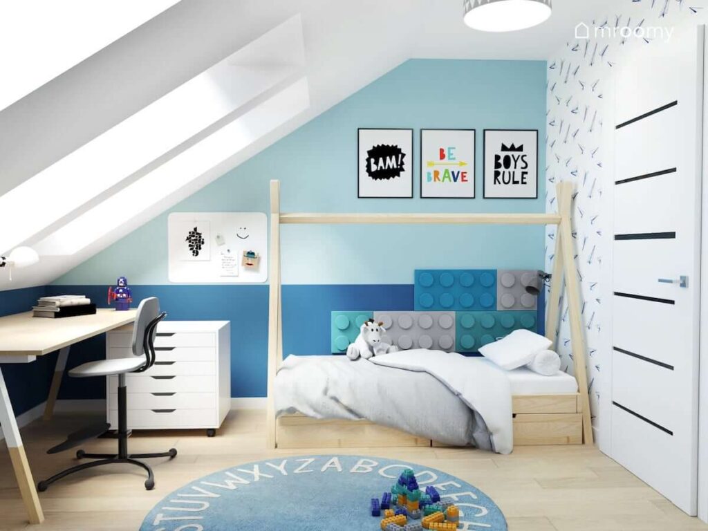 Biało niebieski pokój dla chłopca z drewnianym łóżkiem tipi uzupełnionym miękkimi panelami ściennymi w kształcie klocków lego oraz ozdobnymi plakatami na ścianie i dywanem z alfabetem