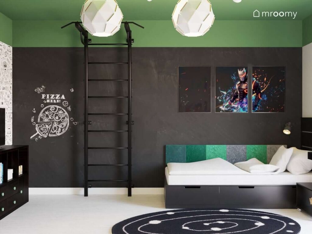 Ściana pomalowana czarną farbą tablicową a na niej drabinka gimnastyczna oraz ozdobny trzyczęściowy plakat a także łóżko uzupełnione panelami ściennymi w różnych kolorach w pokoju dla młodego nastolatka
