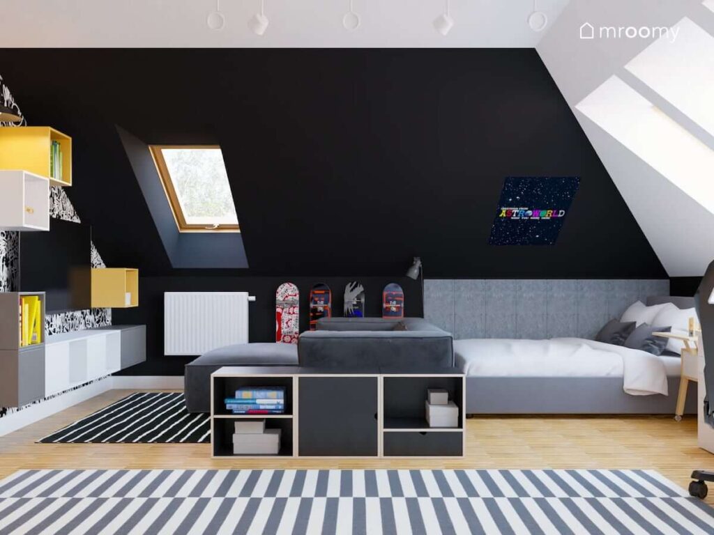 Podwójne łóżko uzupełnione miękkimi panelami ściennymi oraz szara sofa czarny regał ze sklejki i duże dywany w paski w biało czarnym poddaszowym pokoju dla nastolatka