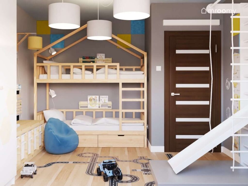 Drewniane łóżko piętrowe w kształcie domku oraz drewniany regał z pojemnikami na zabawki a także żółte i niebieskie panele ścienne a na podłodze naklejka w kształcie jezdni oraz niebieska pufa sako w pokoju dla dwóch chłopców