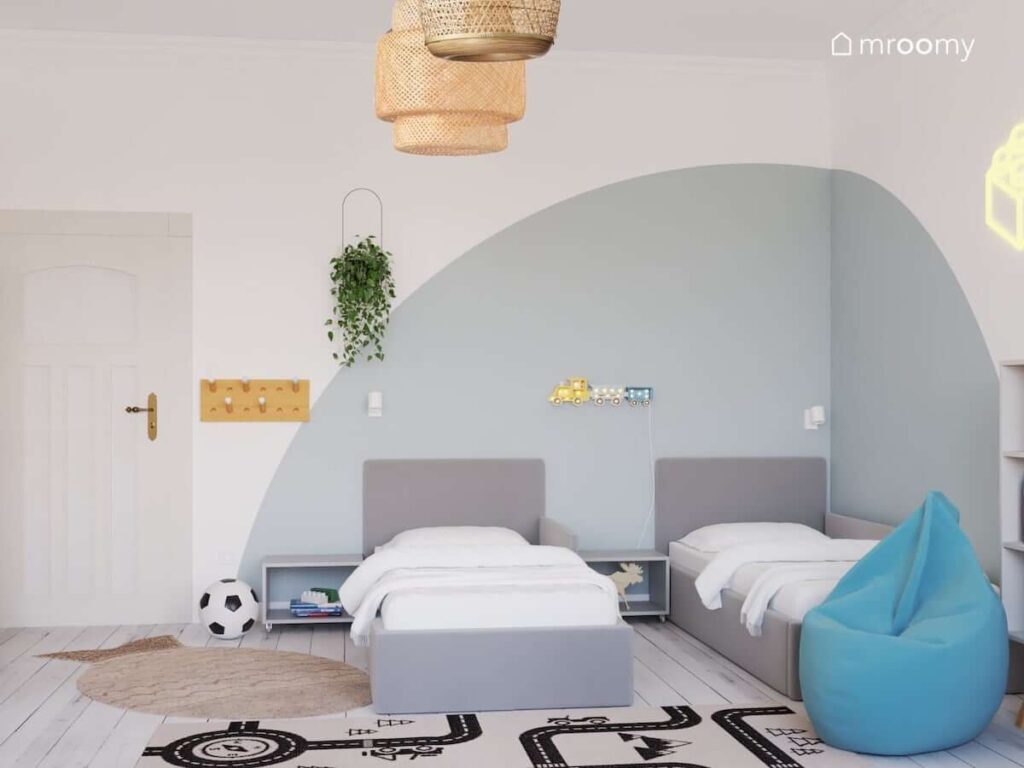 Pokój dla dwóch chłopców z dwoma szarymi łóżkami i stolikami nocnymi na kółkach oraz bambusowymi lampami sufitowymi a na ścianie drewniany wieszak doniczka i lampka nocna w kształcie pociągu
