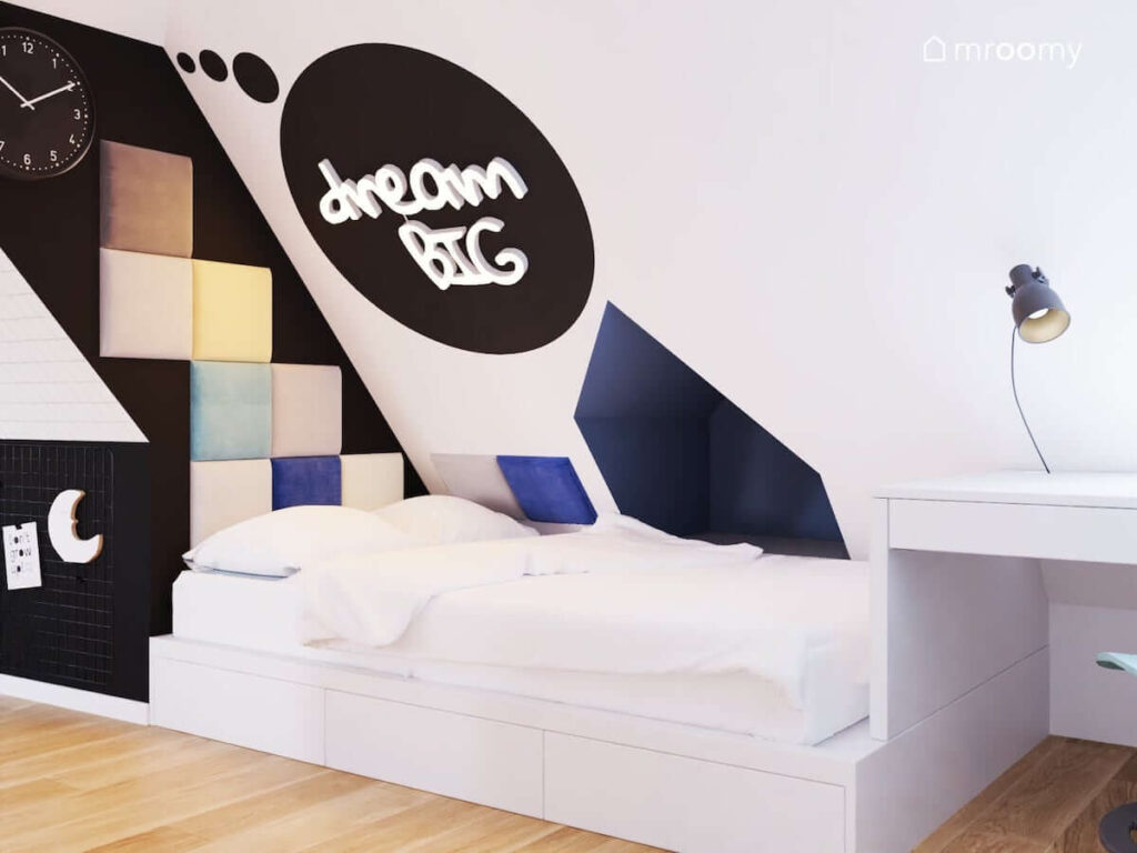 Kącik sypialniany w pokoju dla dwóch chłopców z białym łóżkiem nad którym umieszczono biały ledon w kształcie napisu oraz kolorowe panele ścienne i czarny zegar