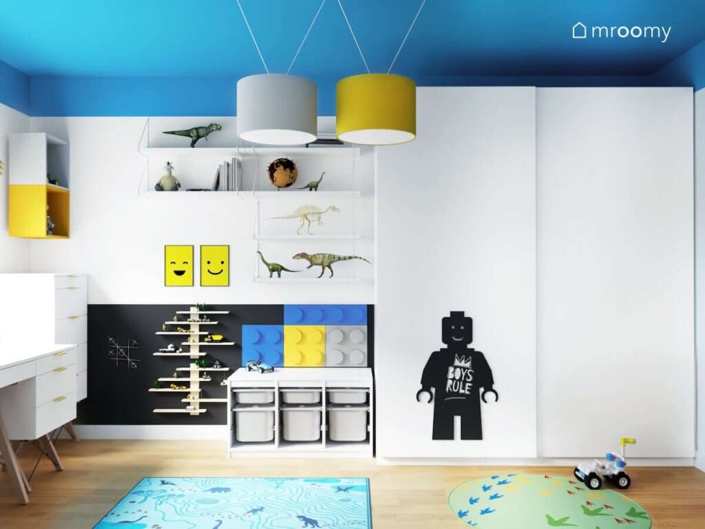 Biało niebieski pokój dla dwóch braci z białą szafą i dywanami w dinozaury oraz z licznymi akcesoriami z motywem Lego takimi jak tablica kredowa ozdobne plakaty i panele ścienne