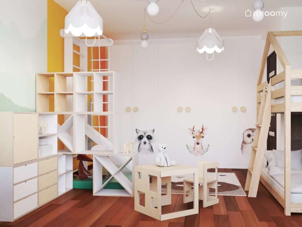 Biało drewniane szafki ze sklejki stolik z krzesełkiem w formie domku oraz duża biała szafa z naklejkami ze zwierzętami leśnymi w pokoju dla dwóch małych sióstr