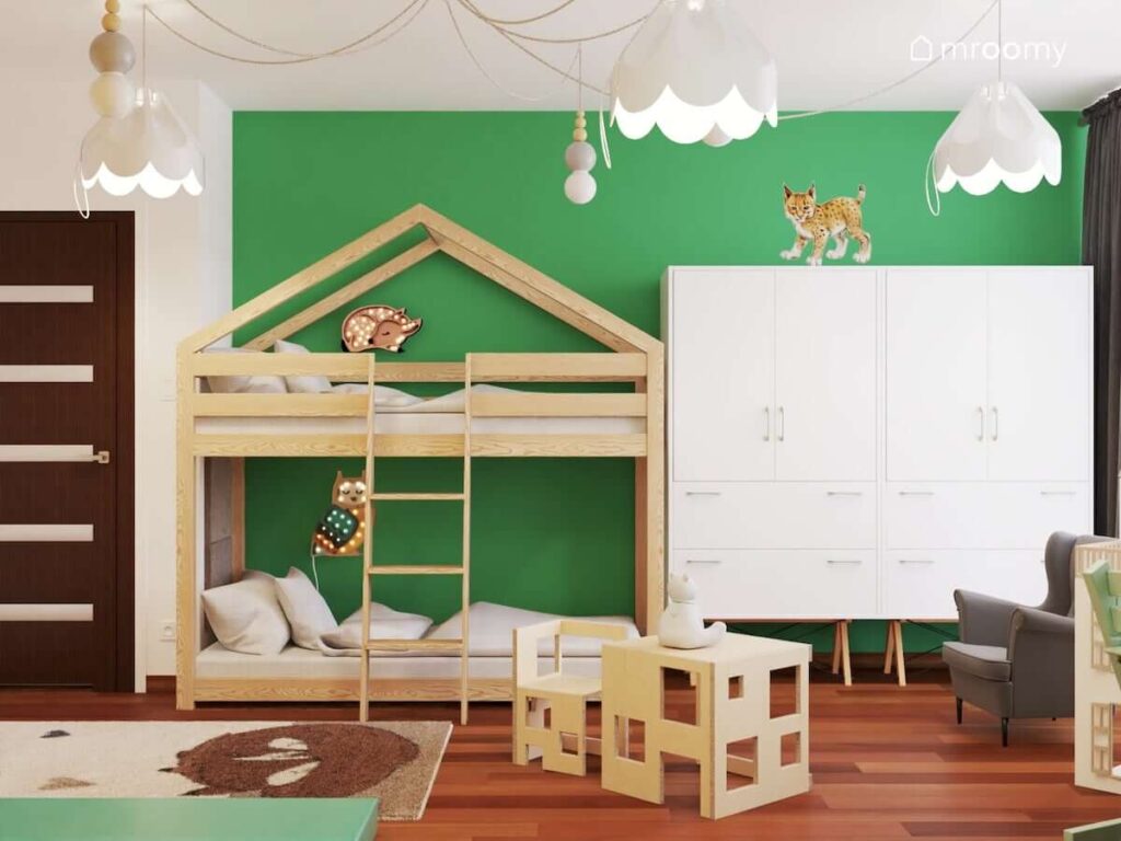 Drewniane łóżko piętrowe w kształcie domku z lampkami nocnymi w kształcie sarny i sowy oraz biała szafa a na suficie ozdobne lampy na rozłożystym zawieszeniu w pokoju dla rodzeństwa z białymi i zielonymi ścianami