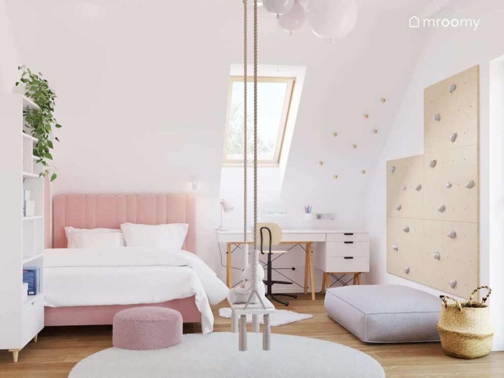 Biały pokój dla kilkulatki z różowym łóżkiem huśtawką wiszącą oraz biurkiem i kontenerkiem na drewnianych nogach oraz z dużym dywanem i wygodnymi pufami na podłodze
